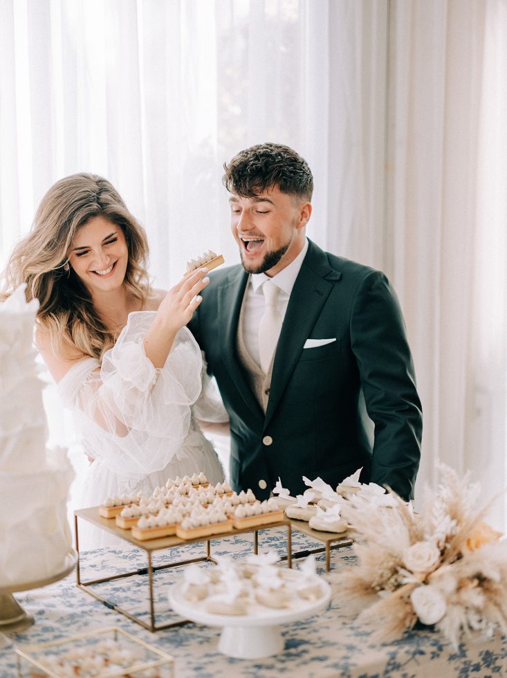 Vrolijk bruidspaar deelt lachend een gebakje bij een feestelijk gedekte desserttafel, in een lichte en romantische setting met zachte kleuren en elegante bloemendecoraties.