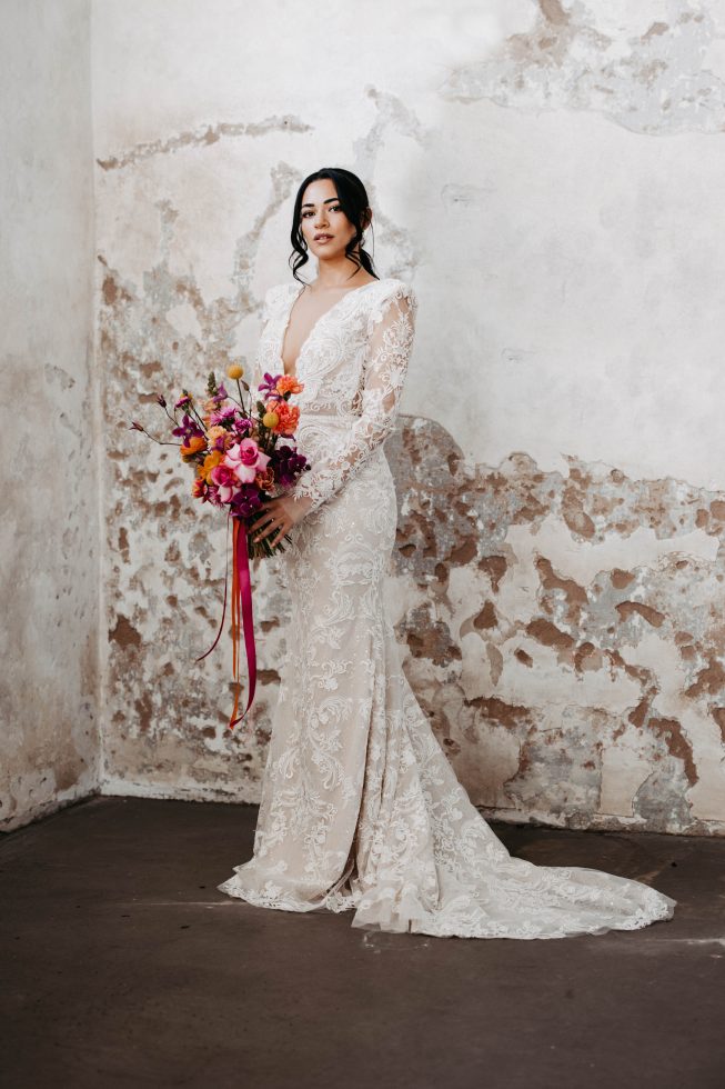 Bruid in een elegante, nauwsluitende kanten trouwjurk met lange mouwen en diepe V-hals, poseert tegen een verweerde muur terwijl ze een kleurrijk boeket met linten vasthoudt.