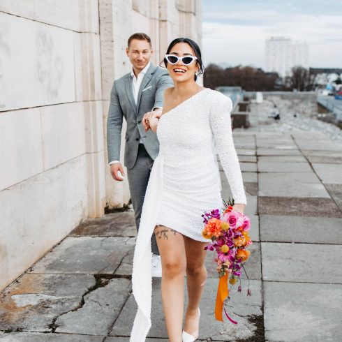 Vrolijke bruid in een korte, moderne trouwjurk en trendy zonnebril leidt lachend haar bruidegom aan de hand over een stedelijke locatie, met een kleurrijk boeket in haar hand.