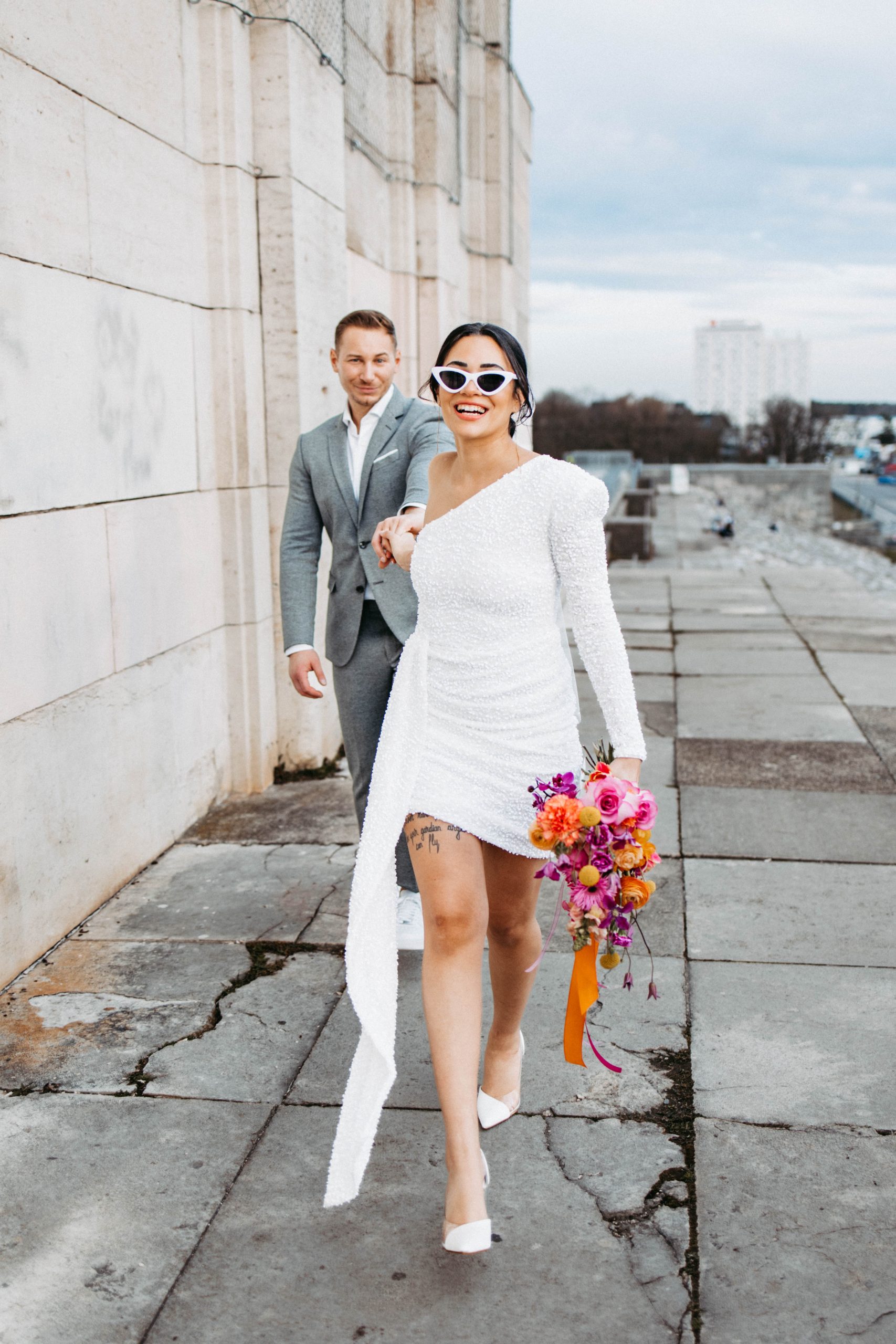 Vrolijke bruid in een korte, moderne trouwjurk en trendy zonnebril leidt lachend haar bruidegom aan de hand over een stedelijke locatie, met een kleurrijk boeket in haar hand.