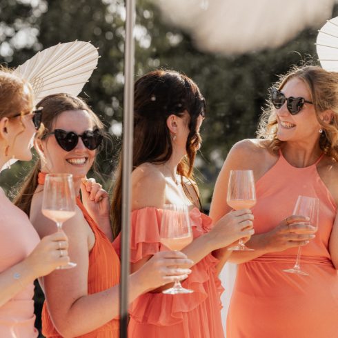 Vier lachende vrouwen in zomerse jurken in tinten koraal en perzik, met zonnebrillen en parasols, proostend met glazen rosékleurige drank tijdens een zonnige buitenbruiloft of feest.