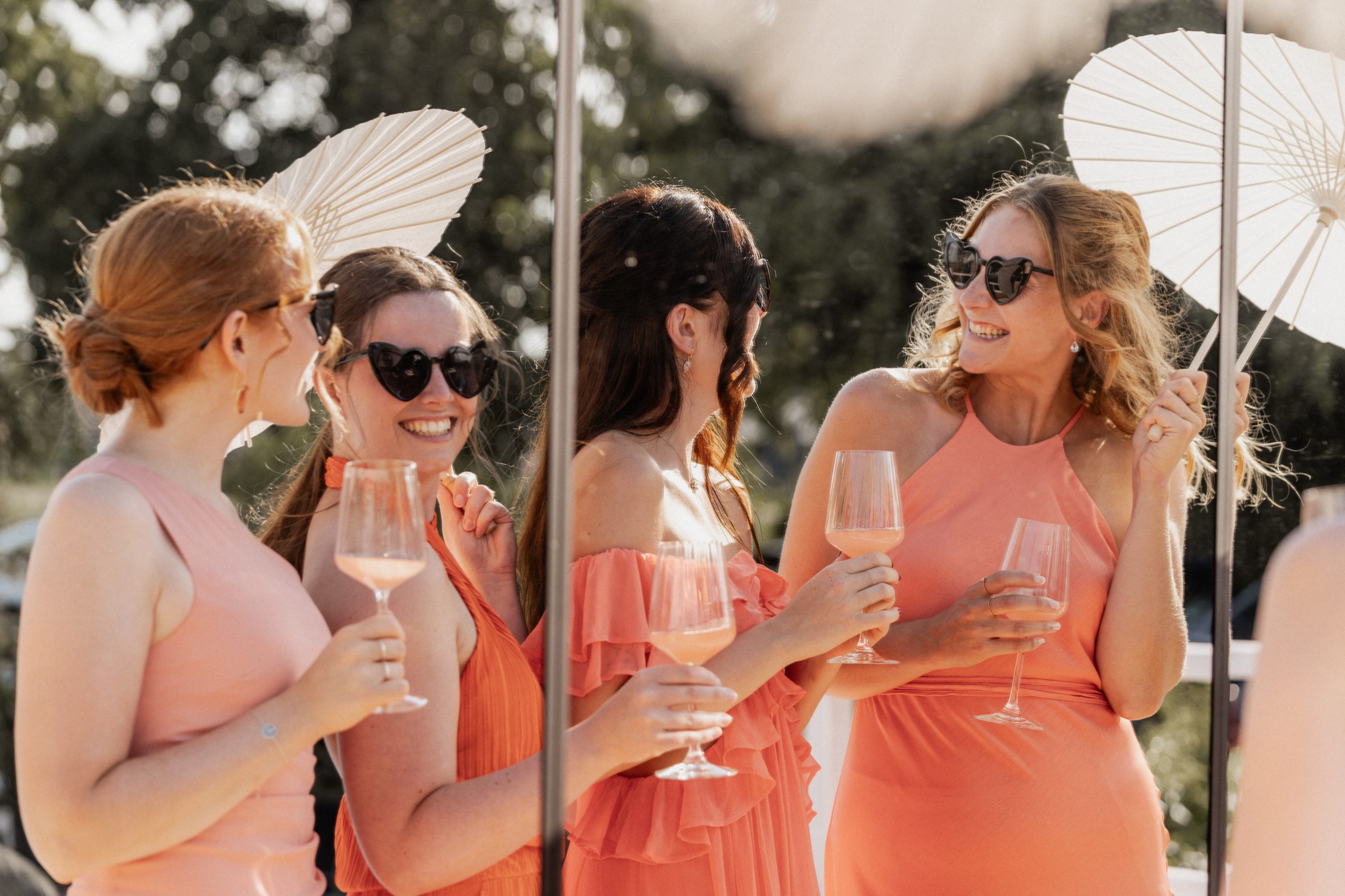 Vier lachende vrouwen in zomerse jurken in tinten koraal en perzik, met zonnebrillen en parasols, proostend met glazen rosékleurige drank tijdens een zonnige buitenbruiloft of feest.