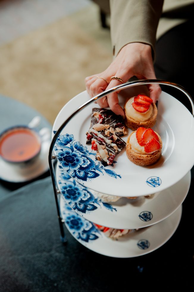 High tea in Hotel Arsenaal in Delft. Twee handen pakken iets van de etagère. Er liggen borden en kopjes bij in Delfts blauwe kleuren.