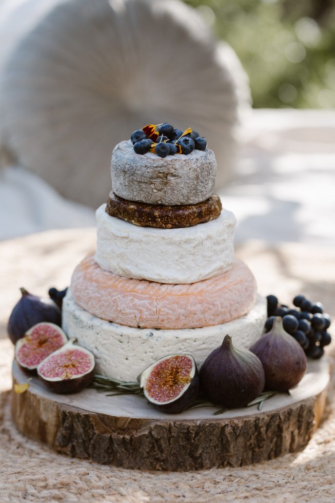 Een gelaagde bruidstaart gemaakt van verschillende soorten kazen, opgemaakt als een traditionele taart, versierd met vijgen, blauwe druiven en bosbessen. De kaastaart staat op een houten plank en wordt omringd door vers fruit, met een zachte, natuurlijke achtergrond.