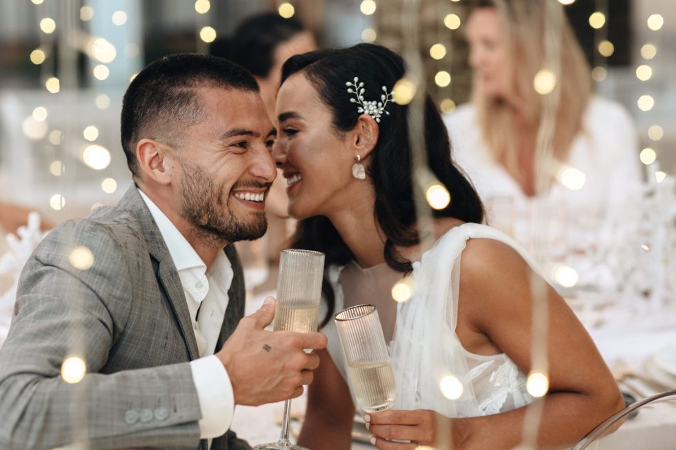 Stralend bruidspaar geniet van een intiem moment tijdens hun feest, proostend met champagneglazen tussen sfeervolle lichtjes in een romantische setting.