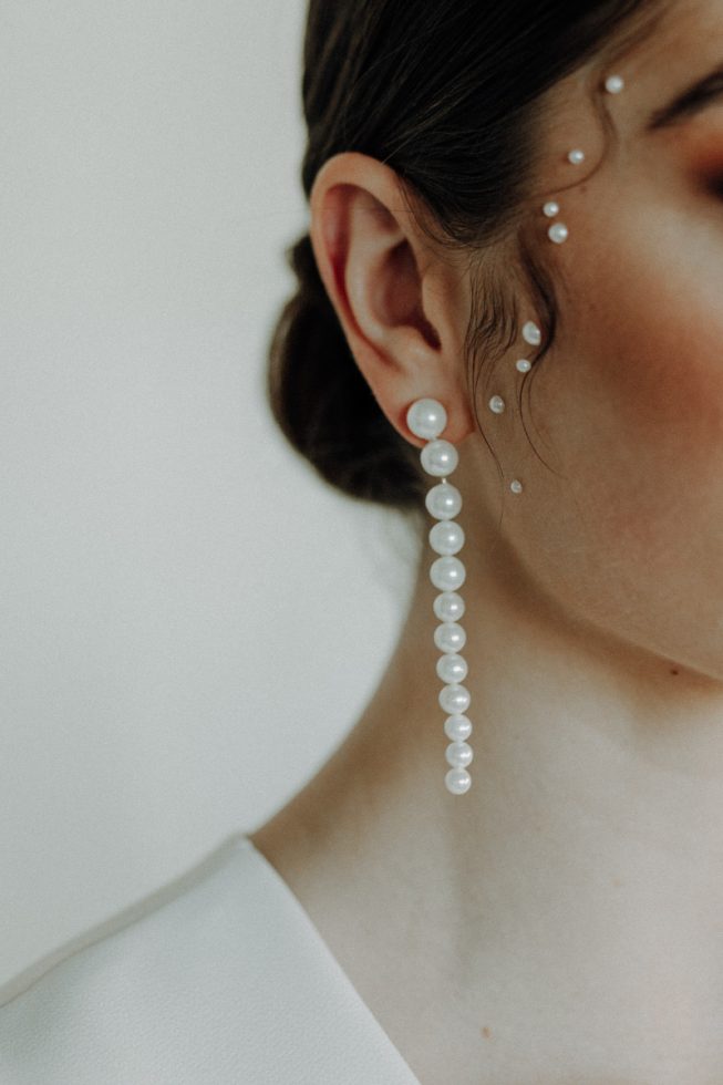 Close-up van een vrouwelijk oor met een lange pareloorbel en kleine parels op de huid langs het gezicht. De huid is licht, het haar is donker en strak naar achteren gestyled.