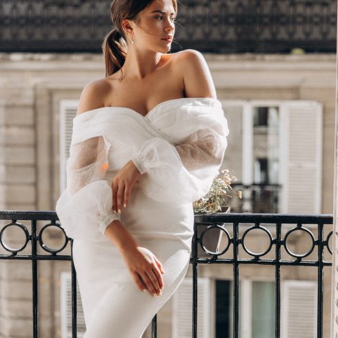 Vrouw in een elegante off-the-shoulder trouwjurk met transparante pofmouwen leunt nonchalant tegen een smeedijzeren balkonhek, met een Parijse gevel op de achtergrond.