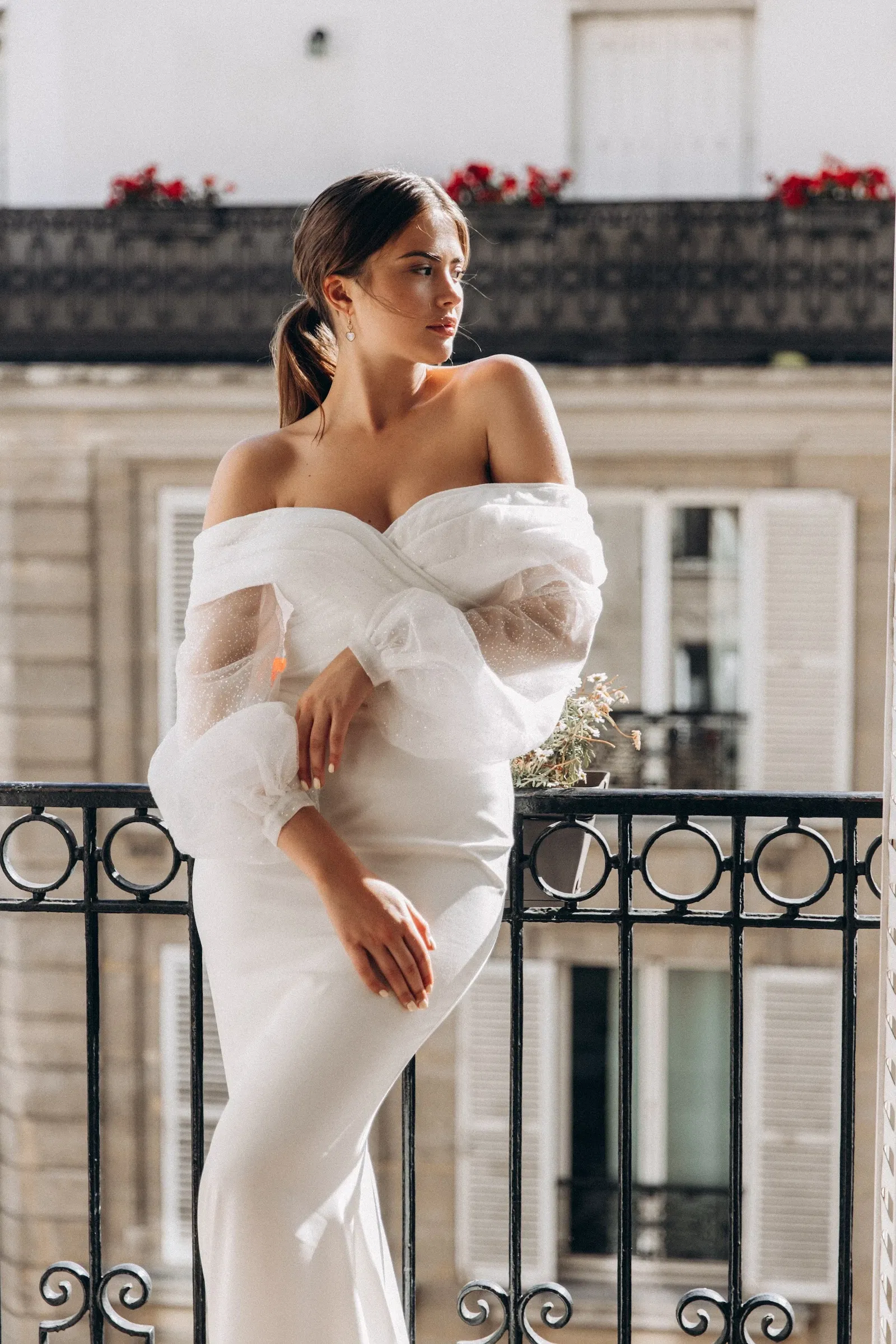 Vrouw in een elegante off-the-shoulder trouwjurk met transparante pofmouwen leunt nonchalant tegen een smeedijzeren balkonhek, met een Parijse gevel op de achtergrond.
