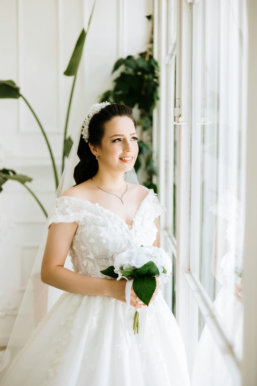 Stralende bruid in een kanten off-shoulder trouwjurk met sluier, die bij een raam staat en een boeket witte bloemen vasthoudt.