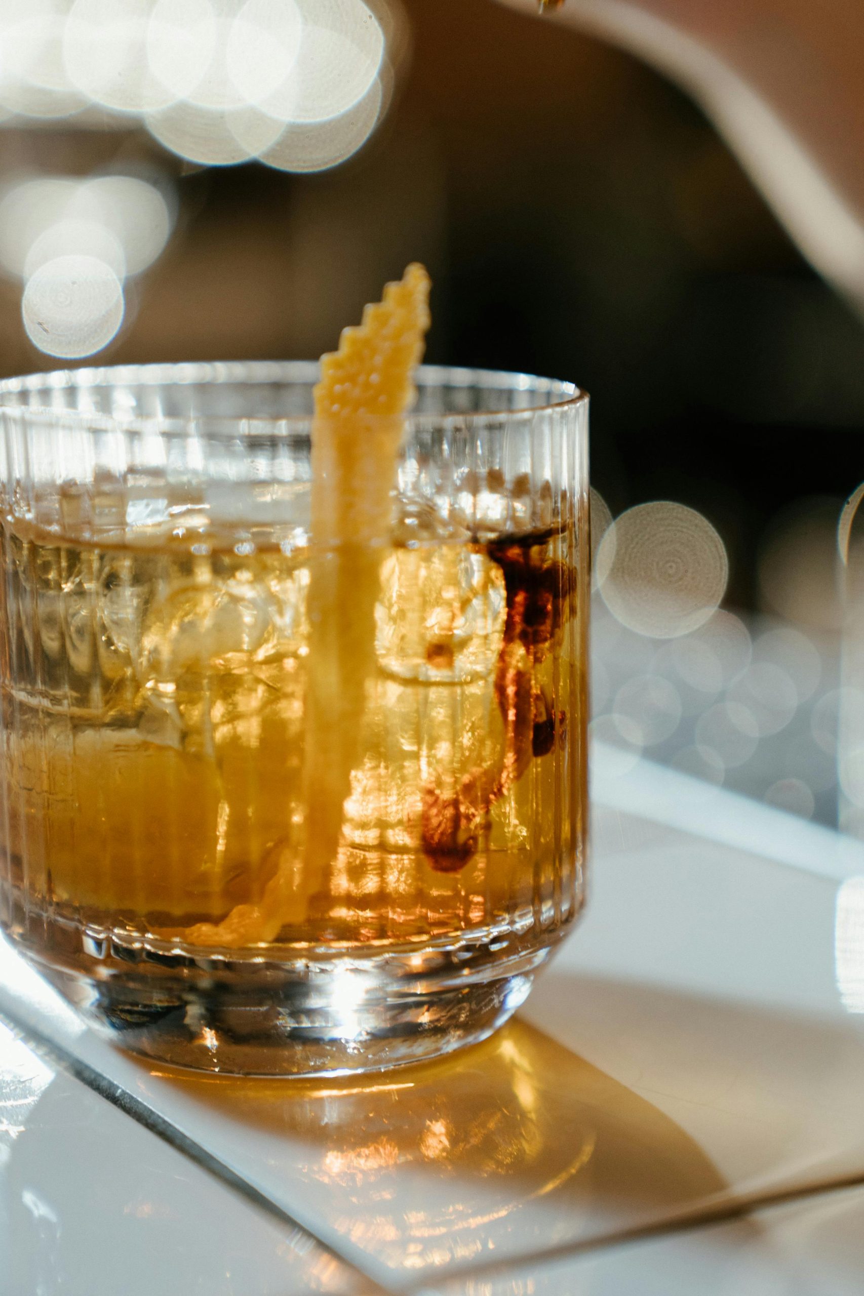 lose-up van een amberkleurige cocktail met ijsblokjes in een geribbeld glas, gegarneerd met een gedraaide citroenschil, met zachte lichtreflecties op de achtergrond.