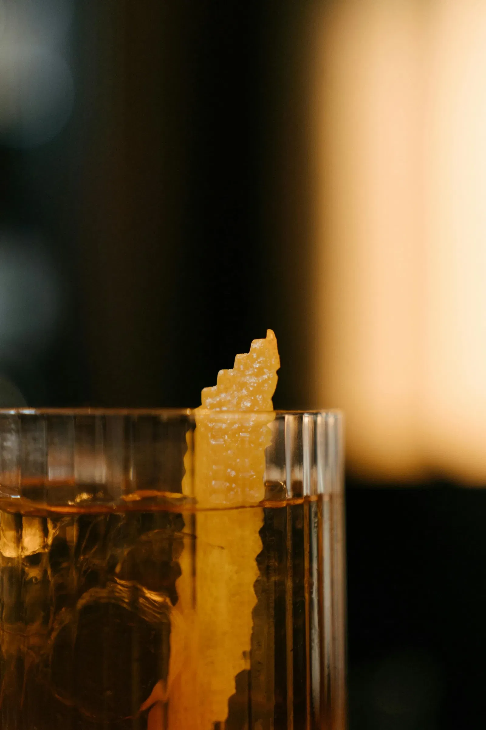 Close-up van een amberkleurige cocktail in een geribbeld glas, gegarneerd met een getande citroenschil, tegen een warme, onscherpe achtergrond.