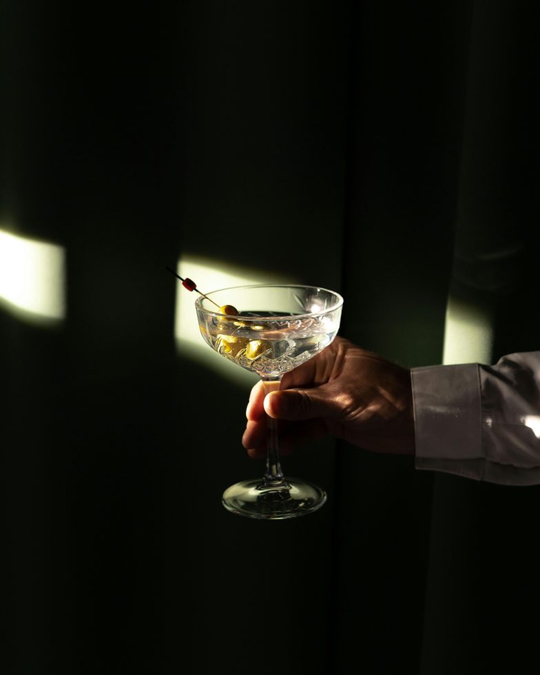 Hand in wit overhemd houdt een stijlvol cocktailglas met een heldere drank en olijven, belicht door dramatisch invallend licht tegen een donkere achtergrond.