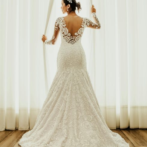 Bruid in een kanten trouwjurk met lange mouwen en open rug, staat voor een helder verlichte gordijnwand en kijkt naar buiten – een moment van rust en verwachting voor de ceremonie.