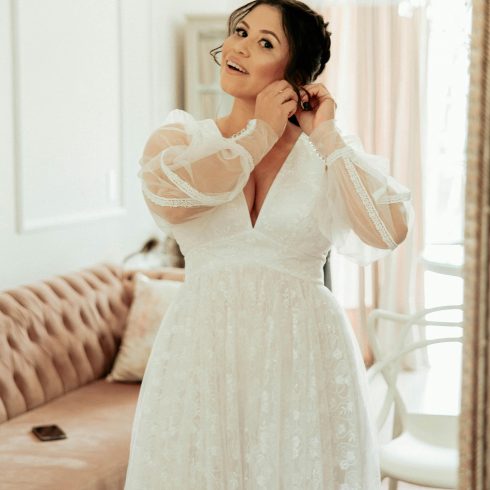 Glimlachende bruid in een romantische trouwjurk met transparante pofmouwen die een oorbel in doet voor een spiegel, in een elegant ingerichte kamer.