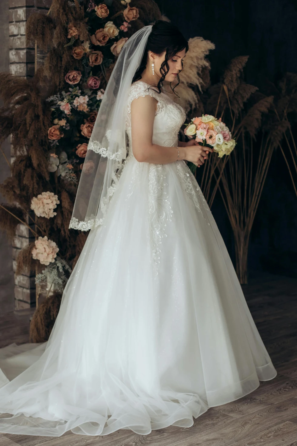 Bruid in een klassieke witte trouwjurk met kant en sluier, houdt een boeket pastelbloemen vast en staat voor een backdrop van droogbloemen en rozen – een intiem moment vol elegantie.