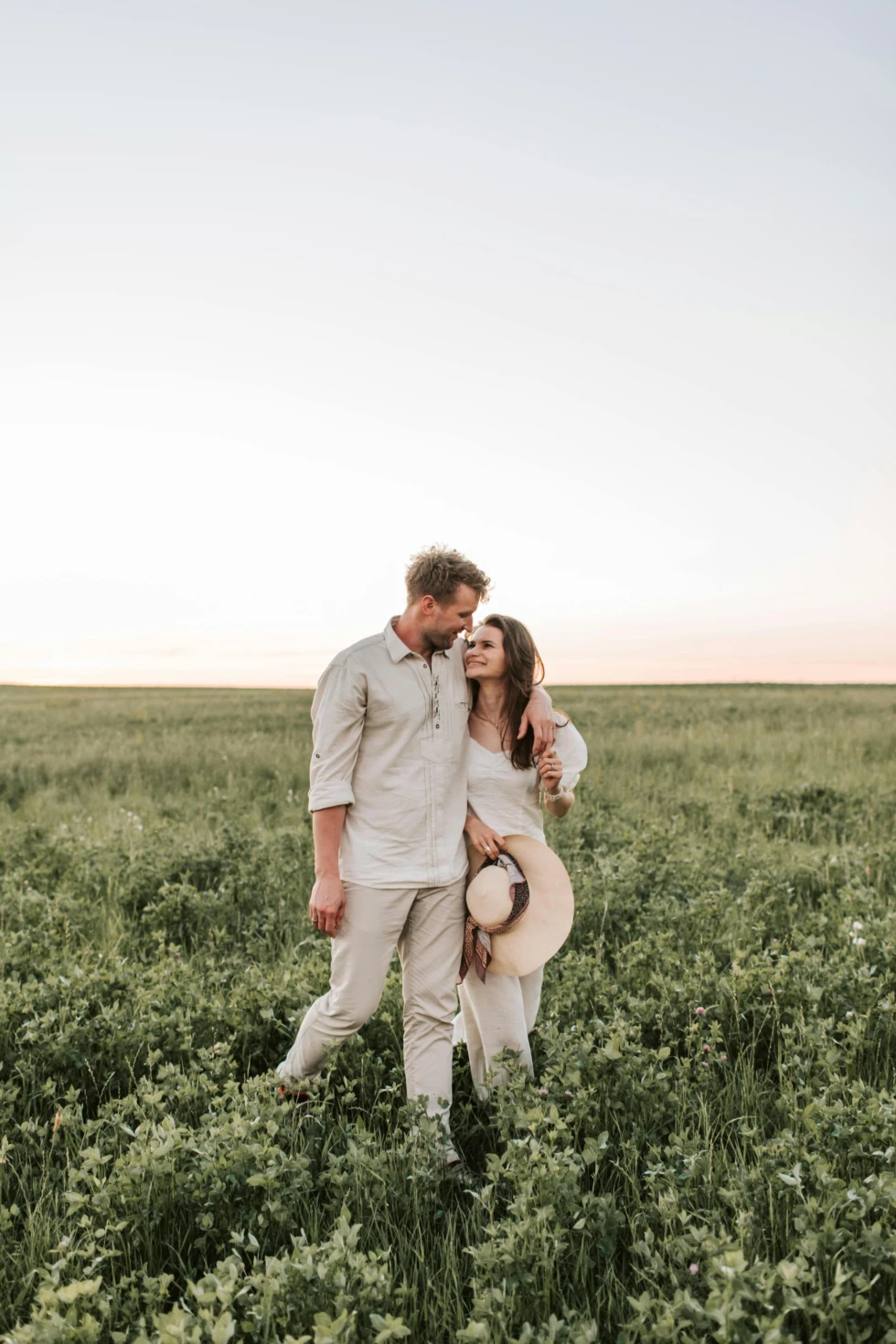 Stel wandelt liefdevol door een groen veld bij zonsondergang, met armen om elkaar heen en een ontspannen, gelukkige uitstraling.