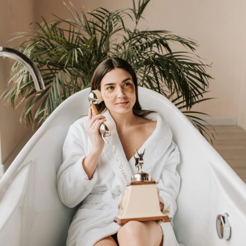 Vrouw in een witte badjas ontspant in een badkuip terwijl ze een vintage telefoon vasthoudt. Ze draagt oogpads en kijkt dromerig voor zich uit, met een groene kamerplant op de achtergrond.