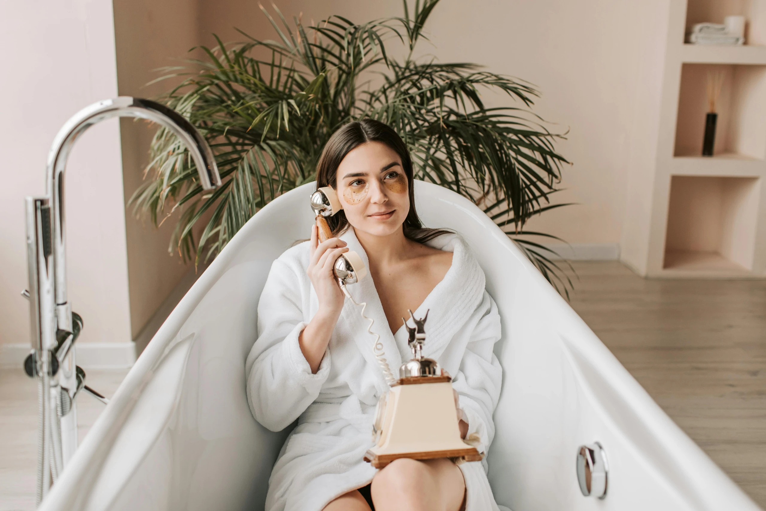 Vrouw in een witte badjas ontspant in een badkuip terwijl ze een vintage telefoon vasthoudt. Ze draagt oogpads en kijkt dromerig voor zich uit, met een groene kamerplant op de achtergrond.