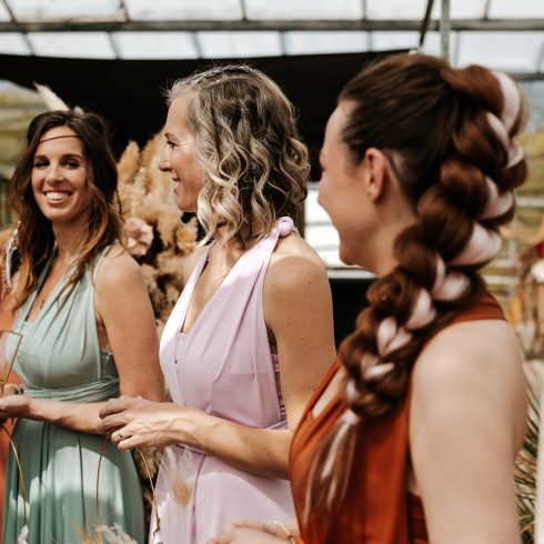 Drie vrouwen gekleed in elegante jurken — mintgroen, lichtroze en roestbruin — staan samen te lachen tijdens een feestelijke gelegenheid in een serre-achtige ruimte. Ze hebben bijzondere kapsels en houden glazen of decoratieve voorwerpen vast. Op de achtergrond zijn pampasgras en andere boho-decoraties te zien, wat een warme en stijlvolle sfeer creëert.