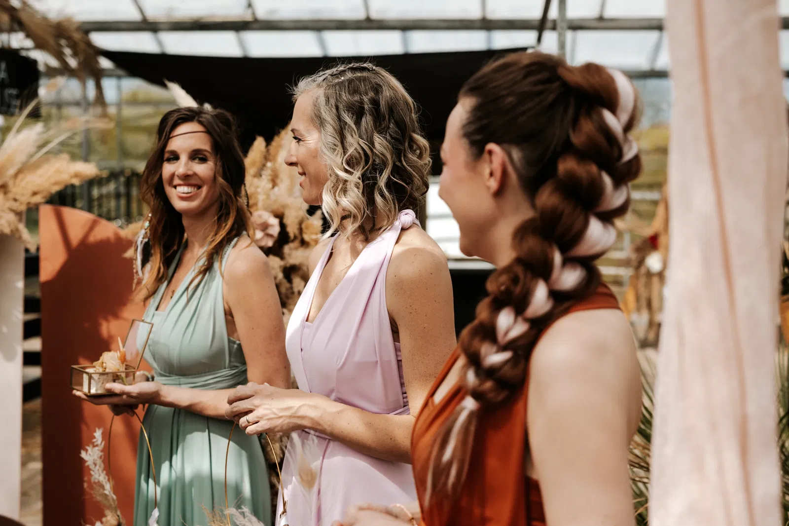 Drie vrouwen gekleed in elegante jurken — mintgroen, lichtroze en roestbruin — staan samen te lachen tijdens een feestelijke gelegenheid in een serre-achtige ruimte. Ze hebben bijzondere kapsels en houden glazen of decoratieve voorwerpen vast. Op de achtergrond zijn pampasgras en andere boho-decoraties te zien, wat een warme en stijlvolle sfeer creëert.