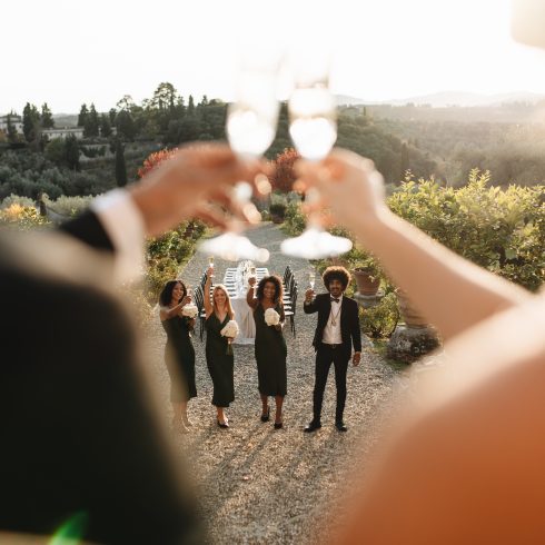 Bruidspaar heft hun glazen tijdens een toostmoment met hun gasten op een sfeervolle buitenbruiloft in Toscane, omringd door groene heuvels en een prachtig gedekte dinertafel.