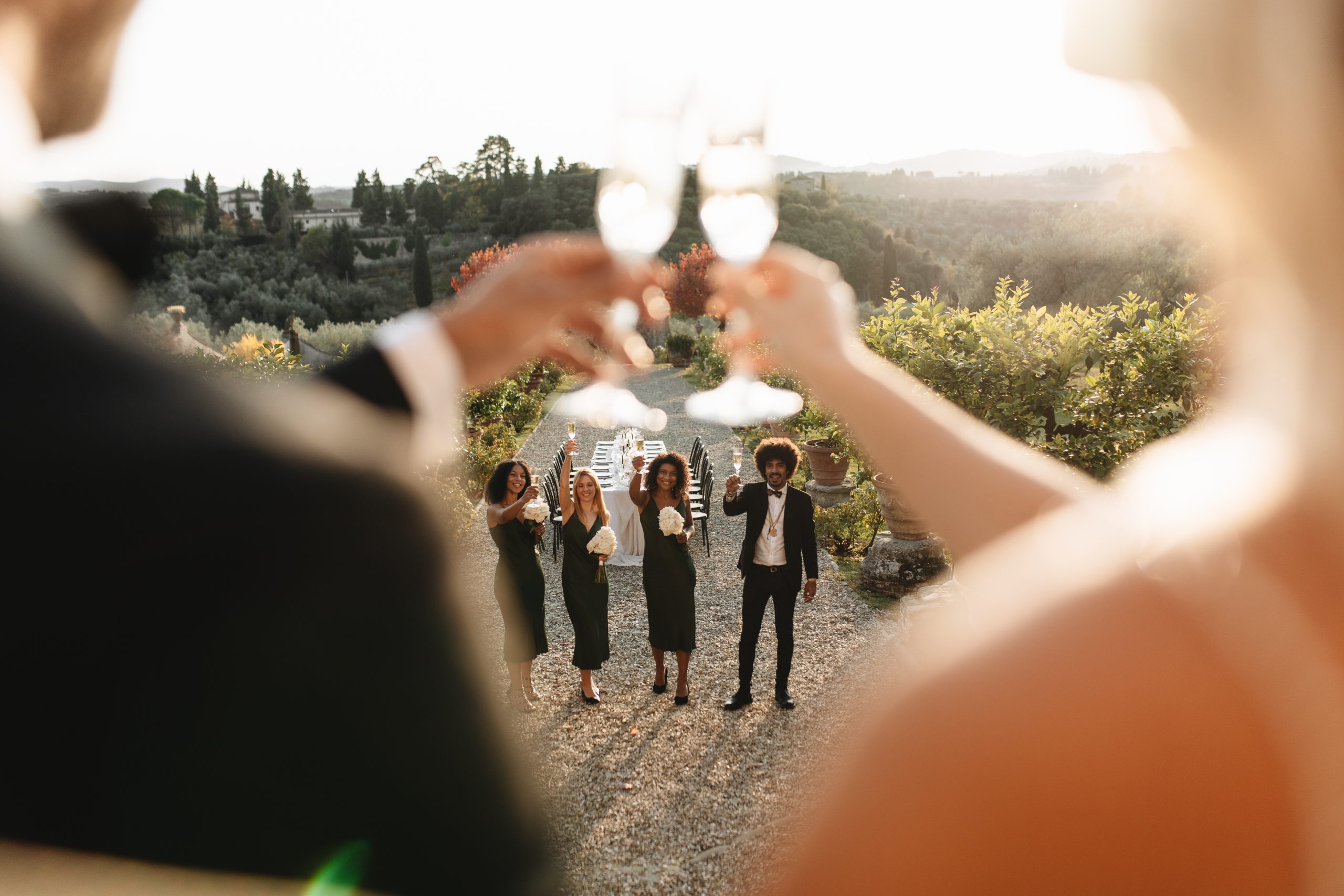 Bruidspaar heft hun glazen tijdens een toostmoment met hun gasten op een sfeervolle buitenbruiloft in Toscane, omringd door groene heuvels en een prachtig gedekte dinertafel.
