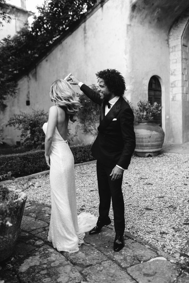 Bruid en bruidegom dansen lachend samen in een sfeervolle, historische buitenlocatie, vastgelegd in een elegante zwart-witfoto die de romantiek en spontaniteit van het moment benadrukt.