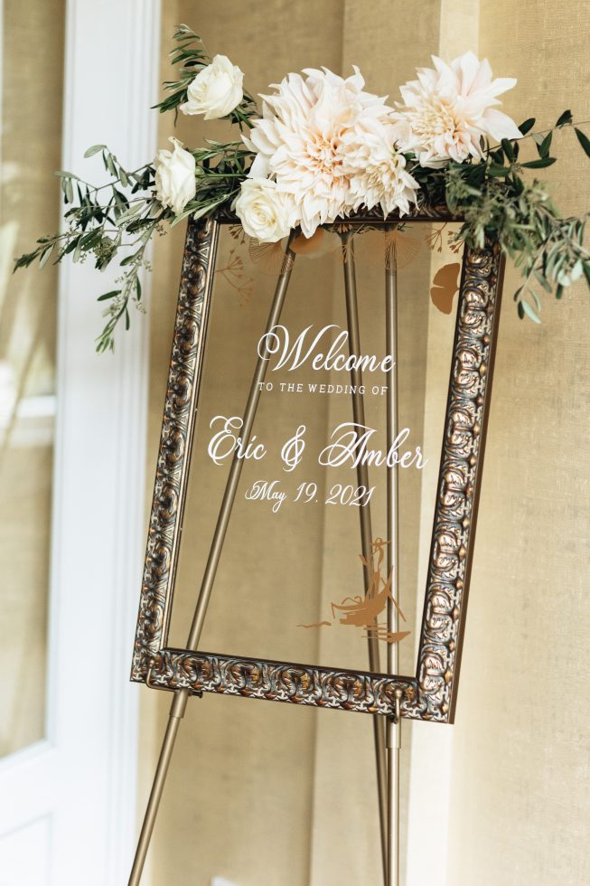 Welkomstbord voor de bruiloft van Eric en Amber op 19 mei 2021, geplaatst in een sierlijke gouden lijst met spiegelachtergrond en versierd met witte bloemen en groen.