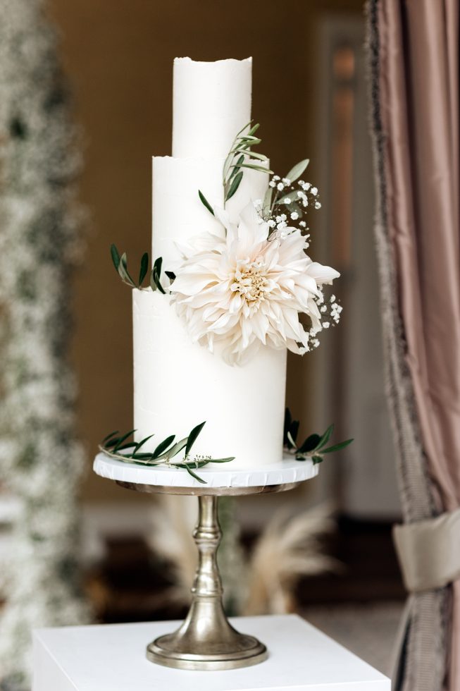 Elegante driedelige bruidstaart met strakke witte afwerking, versierd met een grote crèmekleurige bloem, olijfgroen blad en gipskruid, gepresenteerd op een klassieke zilveren taartstandaard.