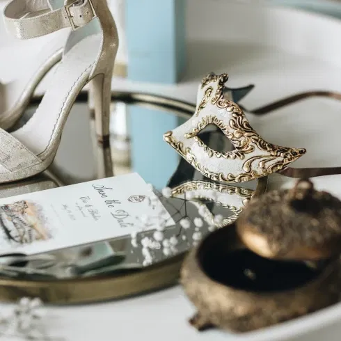 Stijlvolle flatlay met gouden hakken, Venetiaans masker, 'save the date'-kaart, witte roos en vintage sieradendoosje op een spiegelend dienblad – romantische details van een bruiloft met Venetiaans thema.