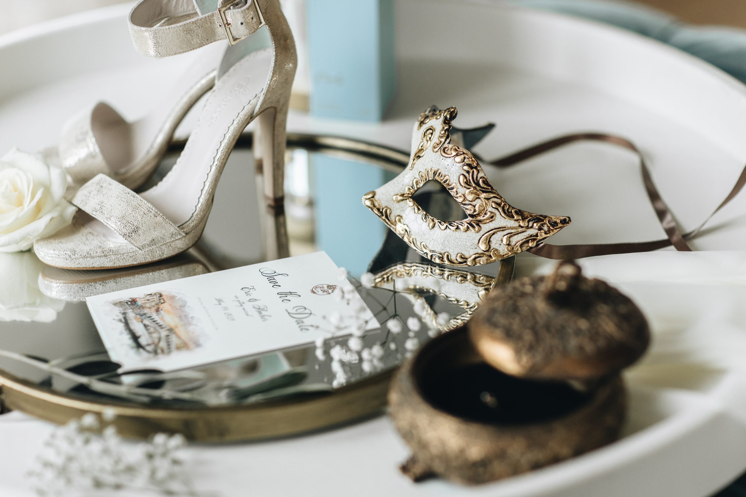 Stijlvolle flatlay met gouden hakken, Venetiaans masker, 'save the date'-kaart, witte roos en vintage sieradendoosje op een spiegelend dienblad – romantische details van een bruiloft met Venetiaans thema.