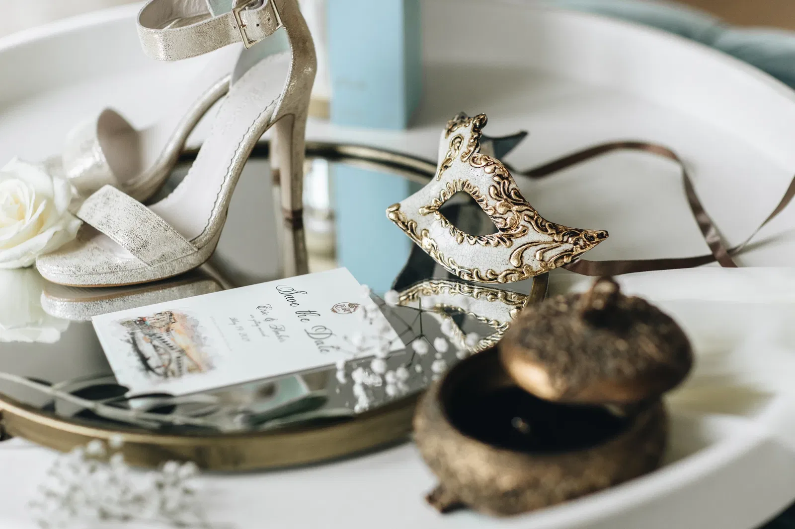 Stijlvolle flatlay met gouden hakken, Venetiaans masker, 'save the date'-kaart, witte roos en vintage sieradendoosje op een spiegelend dienblad โ romantische details van een bruiloft met Venetiaans thema.