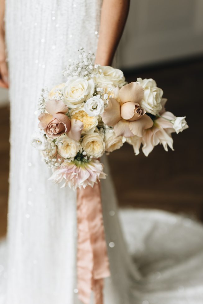 Bruid in glinsterende witte jurk houdt een romantisch boeket vast met crèmekleurige, blush en perzikkleurige rozen, afgewerkt met een lange, zijden lint in zachte perziktint.