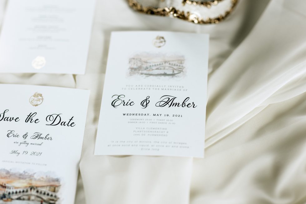 Close-up van stijlvolle trouwkaarten en een 'Save the Date'-kaart voor de bruiloft van Eric en Amber op 19 mei 2021, met een illustratie van Venetië en elegante kalligrafie, liggend op een zijden doek.