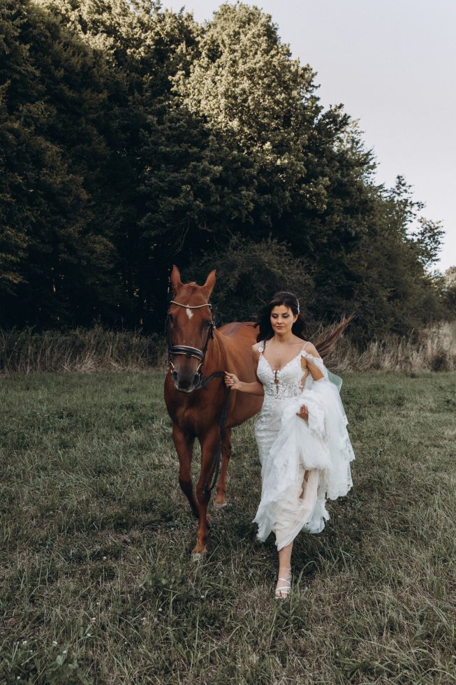 Bruid in een elegante kanten jurk wandelt dromerig over een weide, haar jurk optillend terwijl ze een sierlijk paard aan haar zijde leidt, omringd door groen en rustieke natuur.
