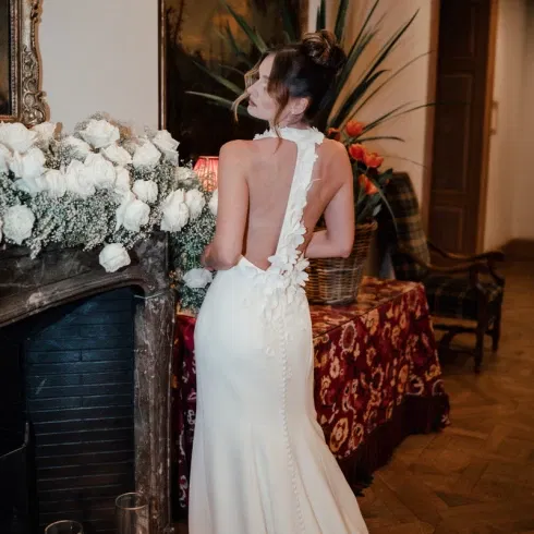 Bruid in elegante, rugloze witte trouwjurk met bloemenapplicaties, staand voor een marmeren schouw versierd met witte rozen en kaarsen.