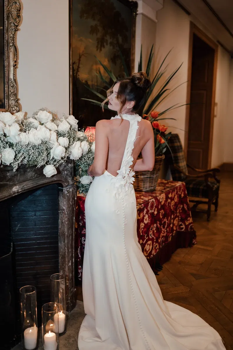 Bruid in elegante, rugloze witte trouwjurk met bloemenapplicaties, staand voor een marmeren schouw versierd met witte rozen en kaarsen.