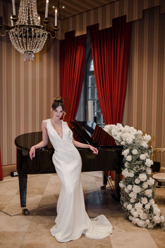 Bruid in elegante witte trouwjurk met diepe V-hals, leunend tegen een zwarte vleugel versierd met witte rozen, in een chique kamer met rode gordijnen en kroonluchter.