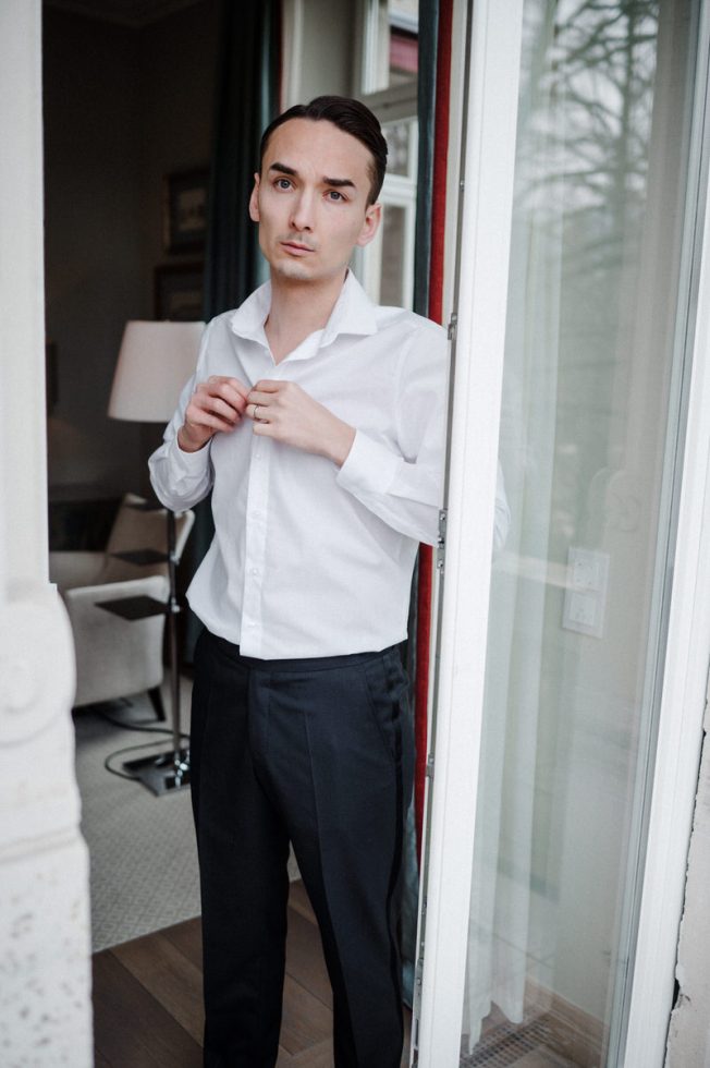 Man in wit overhemd en zwarte pantalon staat bij een open deur terwijl hij zijn overhemd dichtknoopt, klaar om zich verder aan te kleden voor een formele gelegenheid.