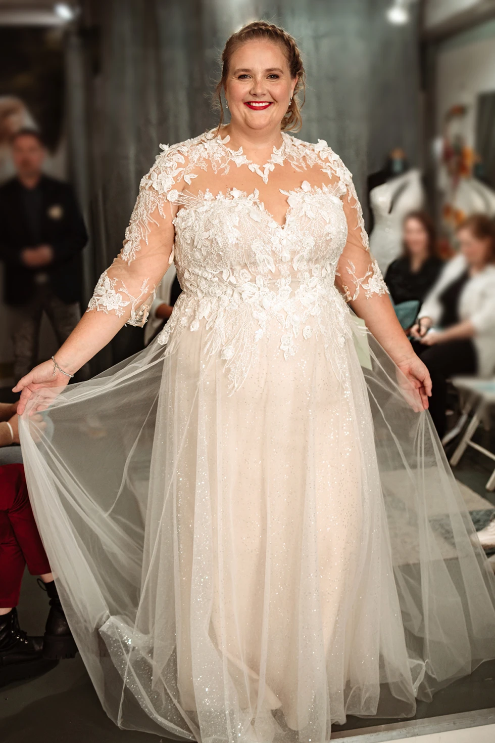 Stralende bruid in een plus size trouwjurk met kant en glitter tijdens een modeshow bij Boetiek de Bruid.