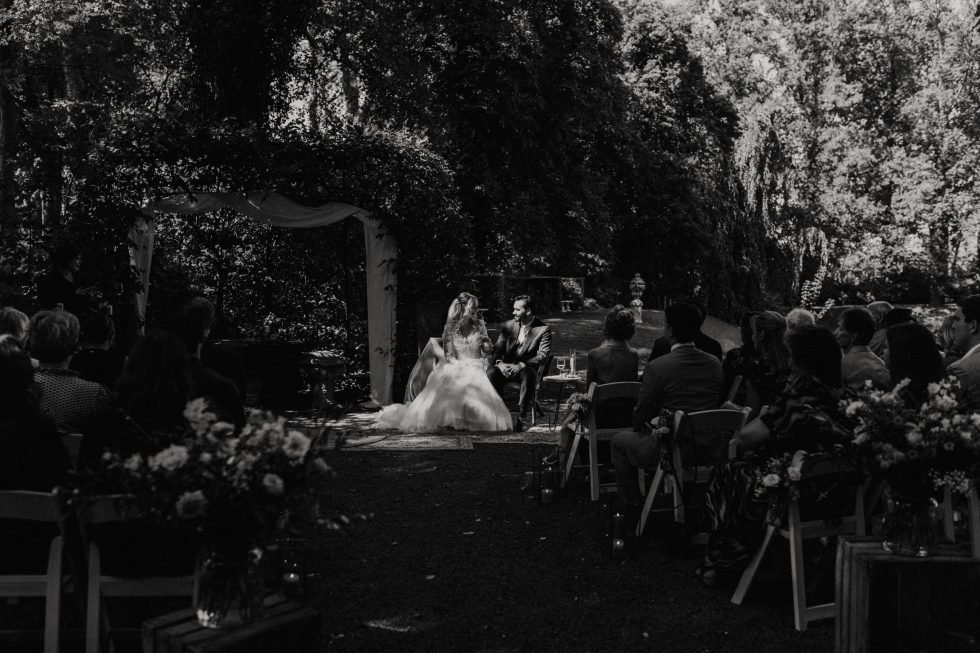 Bruid en bruidegom tijdens romantische buitenbruiloft ceremonie in een bosrijke omgeving, omringd door gasten.
