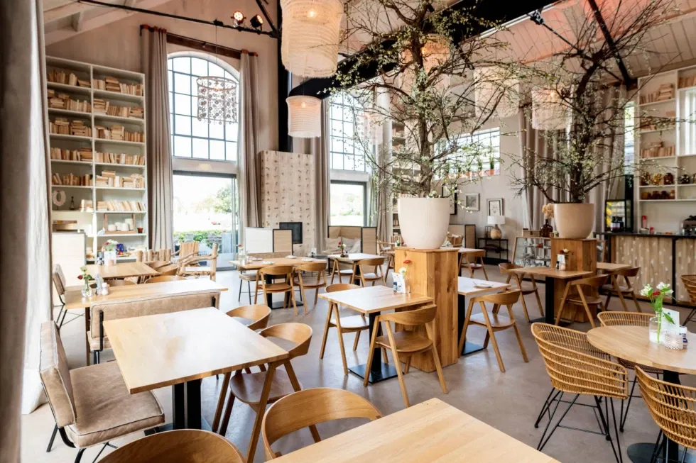 Sfeervolle industriële trouwlocatie met hoge ramen, houten meubels en bloesembomen – warme en stijlvolle setting voor bruiloft of diner.