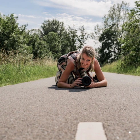 Vrouwelijke fotograaf liggend op het asfalt met camera in de hand – creatieve portretfotografie in de natuur op zomerse dag.