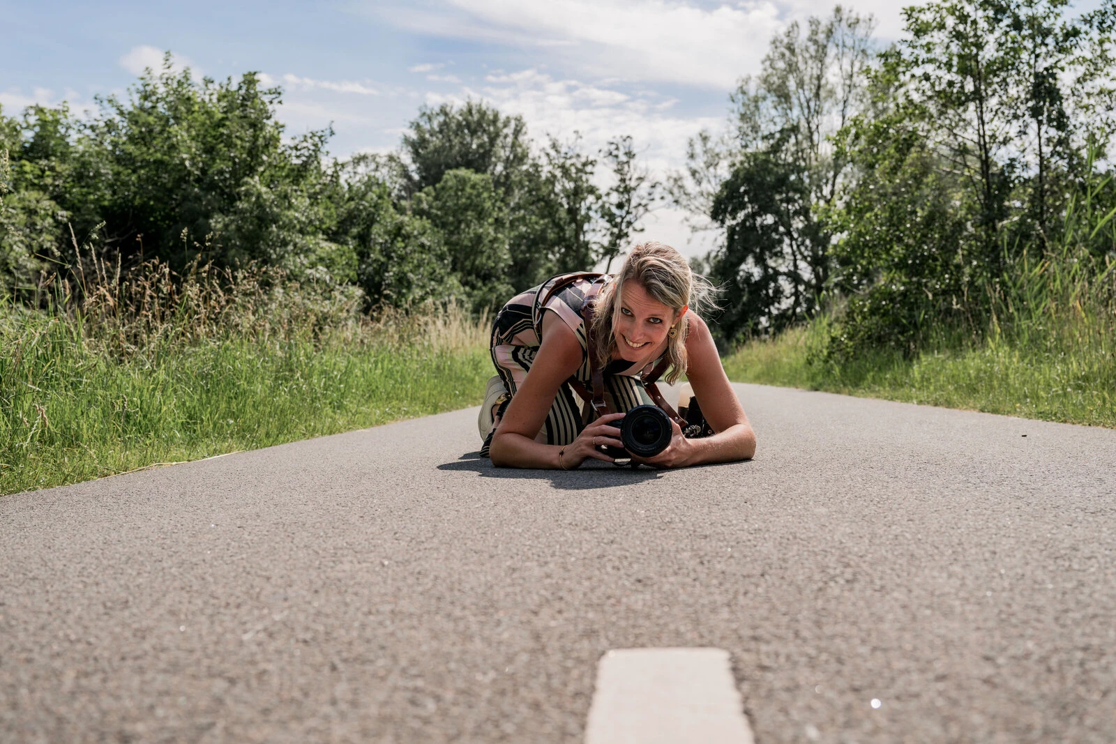 Vrouwelijke fotograaf liggend op het asfalt met camera in de hand – creatieve portretfotografie in de natuur op zomerse dag.