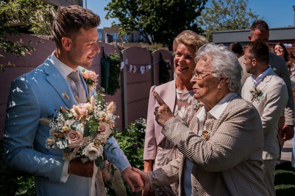 Bruidegom in lichtblauw trouwpak ontvangt emotionele felicitaties van oma tijdens zomerse bruiloft – spontane trouwfoto vol generaties en liefde.