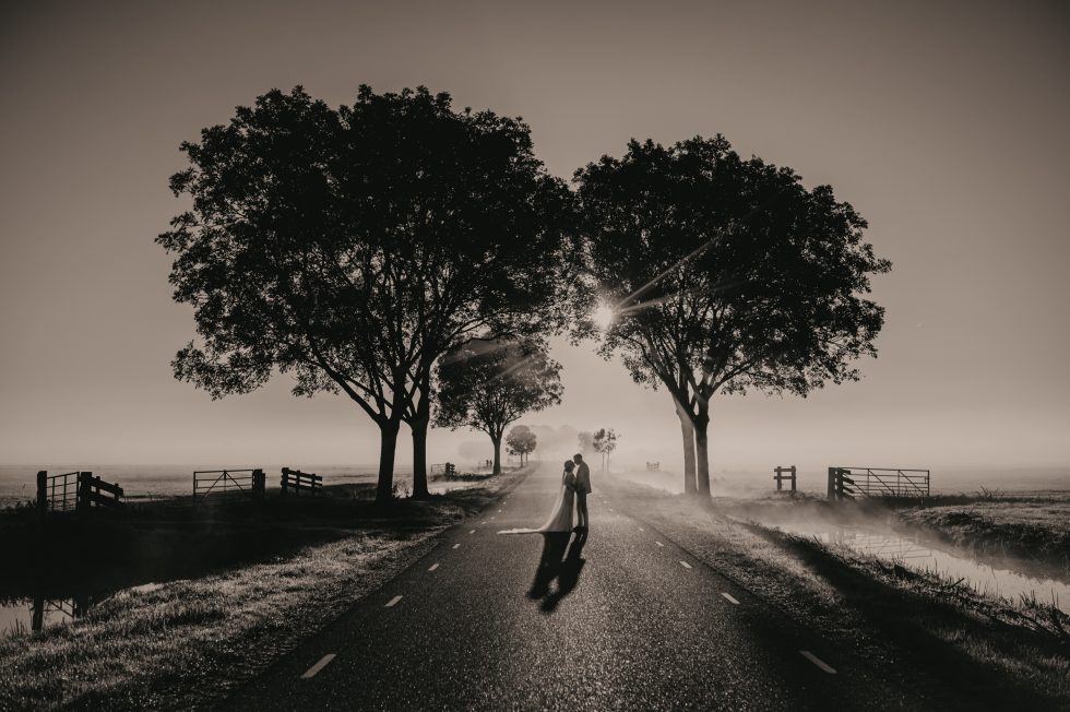 Bruidspaar in de vroege ochtendzon op landweg tussen bomen in landelijke omgeving.