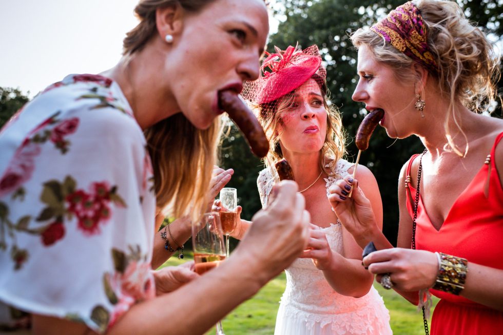 Bruid en gasten eten lachend worstjes op een stok tijdens trouwfeest in de buitenlucht.