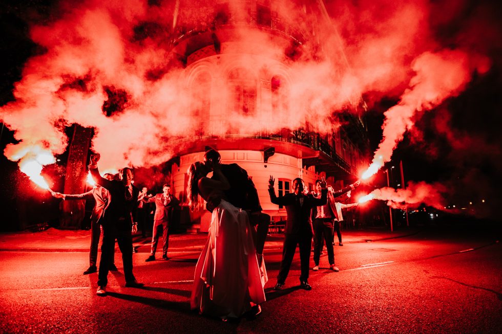 Bruidspaar kust midden op straat tussen vrienden met fakkels in rode rook.
