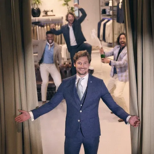 Bruidegom stapt zelfverzekerd uit het pashokje, terwijl zijn vrienden juichend reageren met champagne in een stijlvolle mannenmodewinkel.
