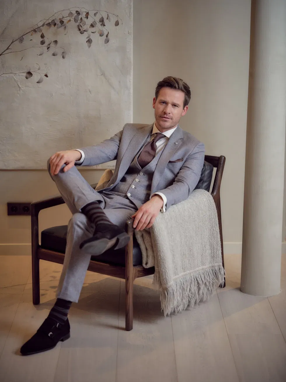 Stijlvol geklede man in een lichtgrijs trouwpak poseert ontspannen zittend op een designstoel in een luxe interieur.
